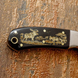 The handle is crafted from polished bull horn, presenting deep dark tones that contrast beautifully with the golden wildlife engraving across its surface.