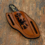 The handcrafted brown leather sheath continues the wilderness theme with a deeply engraved moose standing beside tall pine trees, capturing the atmosphere of a northern forest landscape.