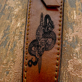 The artwork depicts a ram skull with long curling horns pierced by a sword, while a serpent coils around the blade, creating a dramatic and powerful mythic motif.