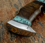 Close-up of a custom knife handle featuring a polished silver-colored bolster, a turquoise inlay with black veining, and a dark brown wood handle