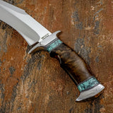 A custom fixed-blade knife with a silver blade, polished steel guard and pommel, and a dark brown wood handle inlaid with turquoise-colored stone. 