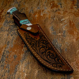 A handcrafted knife with turquoise inlay and a dark wood handle, partially tucked into a brown leather sheath featuring an engraved floral scroll pattern