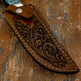 A close-up of a handcrafted brown leather knife sheath with intricate floral tooling and contrast stitching