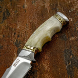 A close-up of a fixed-blade knife with a polished silver blade, a cream-colored bone handle and gold-toned geometric metal filework