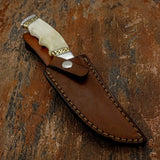 "Sandstone" A fixed-blade knife with an off-white bone handle and gold-toned guard, resting inside a brown leather sheath 