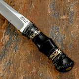 A close-up of a knife handle shaped from polished black bull horn with intricate hand-worked contours, and decorative brass fileworks. These fine details give the dark handle a distinctive ornamental character without overwhelming its slender form.
