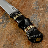 A close-up of a knife handle The handle is the centrepiece of the design, shaped from polished black bull horn with intricate hand-worked contours, and decorative brass fileworks. These fine details give the dark handle a distinctive ornamental character without overwhelming its slender form.
