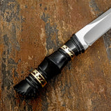 A close-up of a knife handle shaped from polished black bull horn with intricate hand-worked contours, and decorative brass fileworks. These fine details give the dark handle a distinctive ornamental character without overwhelming its slender form.