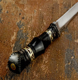 A close-up of a decorative knife or knife handle and partial blade resting on a textured brown stone surface. The handle is glossy black with ornate gold geometric bands and a sculpted black pommel, while the blade is silver-toned metal.