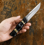 A hand holding a fixed-blade custom knife with a satin-finished D2 silver blade and a bull horn handle featuring brass geometric accents