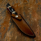A fixed-blade knife with a dark handle and decorative gold-toned bands, partially tucked into a brown leather sheath with visible stitching and a snap closure