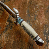 Spine of the custom bowie Canyon Scroll, Intricate black scrollwork across the camel bone forms the centrepiece of the handle, complemented by carefully arranged gemstone sections, contrasting spacers and sculpted metalwork. The matching scroll-patterned leather sheath completes the presentation.
