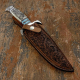 A handcrafted brown leather sheath accompanies the knife. Its face carries extensive flowing scroll decoration that visually complements the engraved handle, together with contrasting stitching and a secure retention strap.
