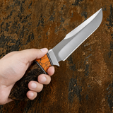 A hand holding a fixed-blade knife with a satin-finished D2 steel blade and a handle made of carved wood and bright orange Gemstone inlay