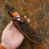 A hand holds a fixed-blade knife with an orange Gemstone and wood handle, resting on a dark brown leather sheath with floral tooling