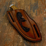 The handcrafted brown leather sheath features a deeply engraved wilderness scene of two wolves standing on rocky ground, howling toward the sky.