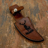 The handcrafted leather sheath continues the wilderness theme with a detailed engraving of a standing elk with broad antlers, depicted in a natural outdoor setting.