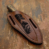 The handcrafted brown leather sheath features a deeply engraved wilderness scene depicting a wolf howling while another wolf stands alert above on rugged terrain.